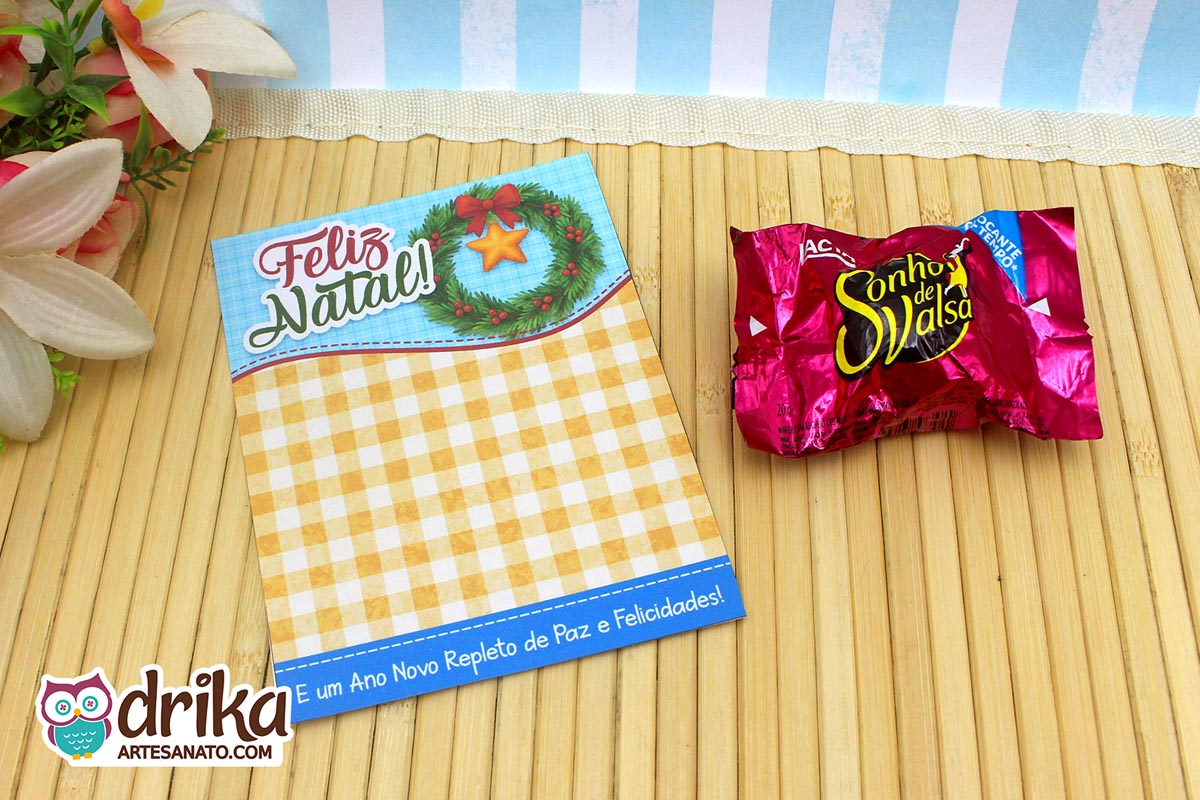 Cartão porta-bombom de Natal com guirlanda, laço vermelho e bombom Sonho de Valsa ao lado, sobre esteira de bambu.