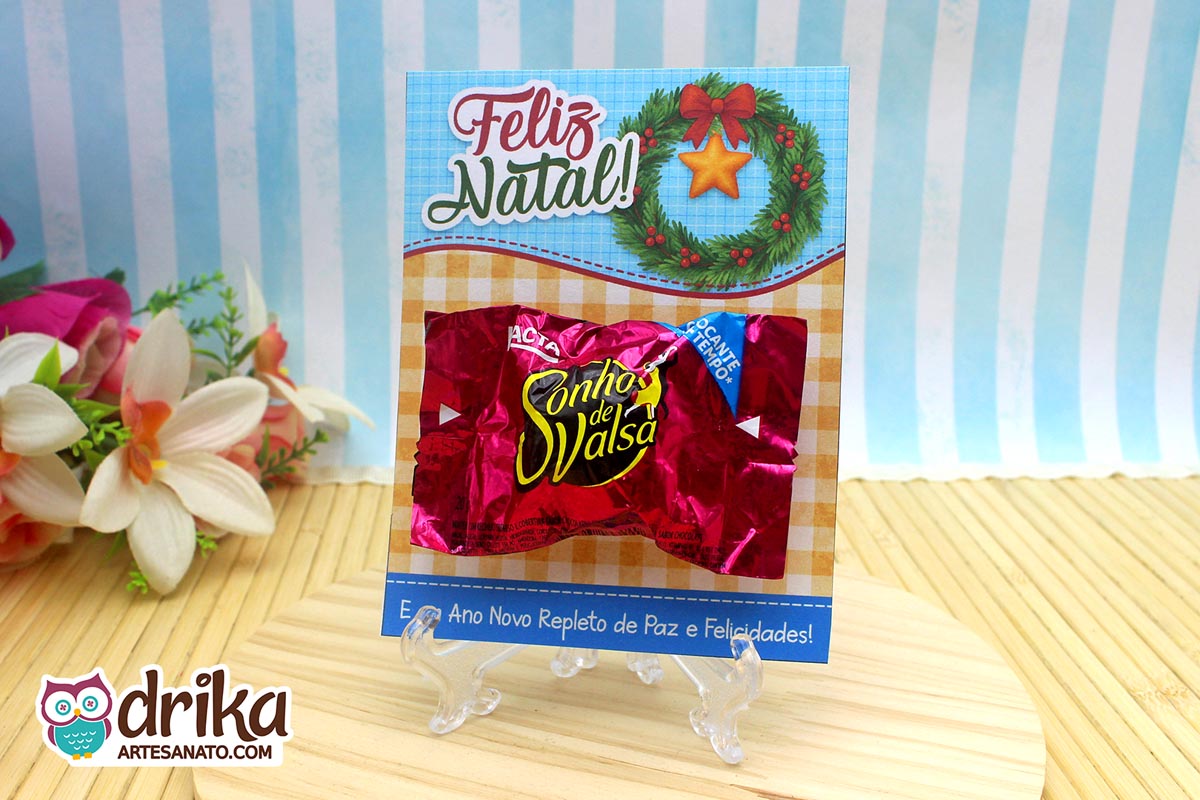 Cartão porta-bombom de Natal com guirlanda, laço vermelho e bombom Sonho de Valsa afixado, em base transparente.