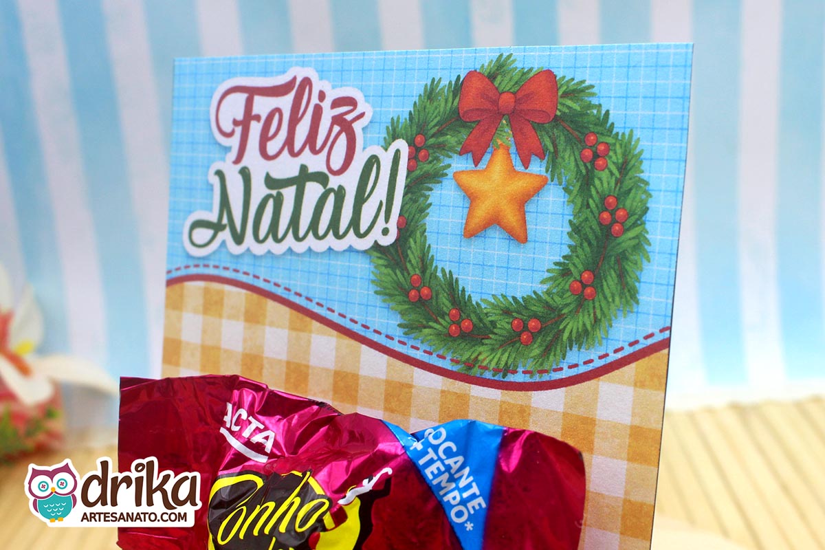 Detalhe do cartão de Natal com guirlanda verde, laço vermelho, estrela dourada e a frase "Feliz Natal!".