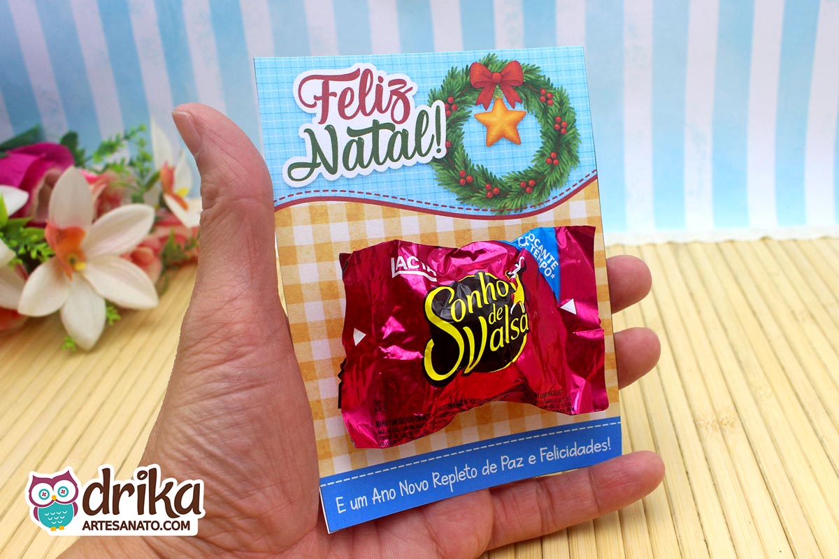 Mão segurando cartão porta-bombom de Natal com bombom Sonho de Valsa, com fundo xadrez e mensagem de Ano Novo.