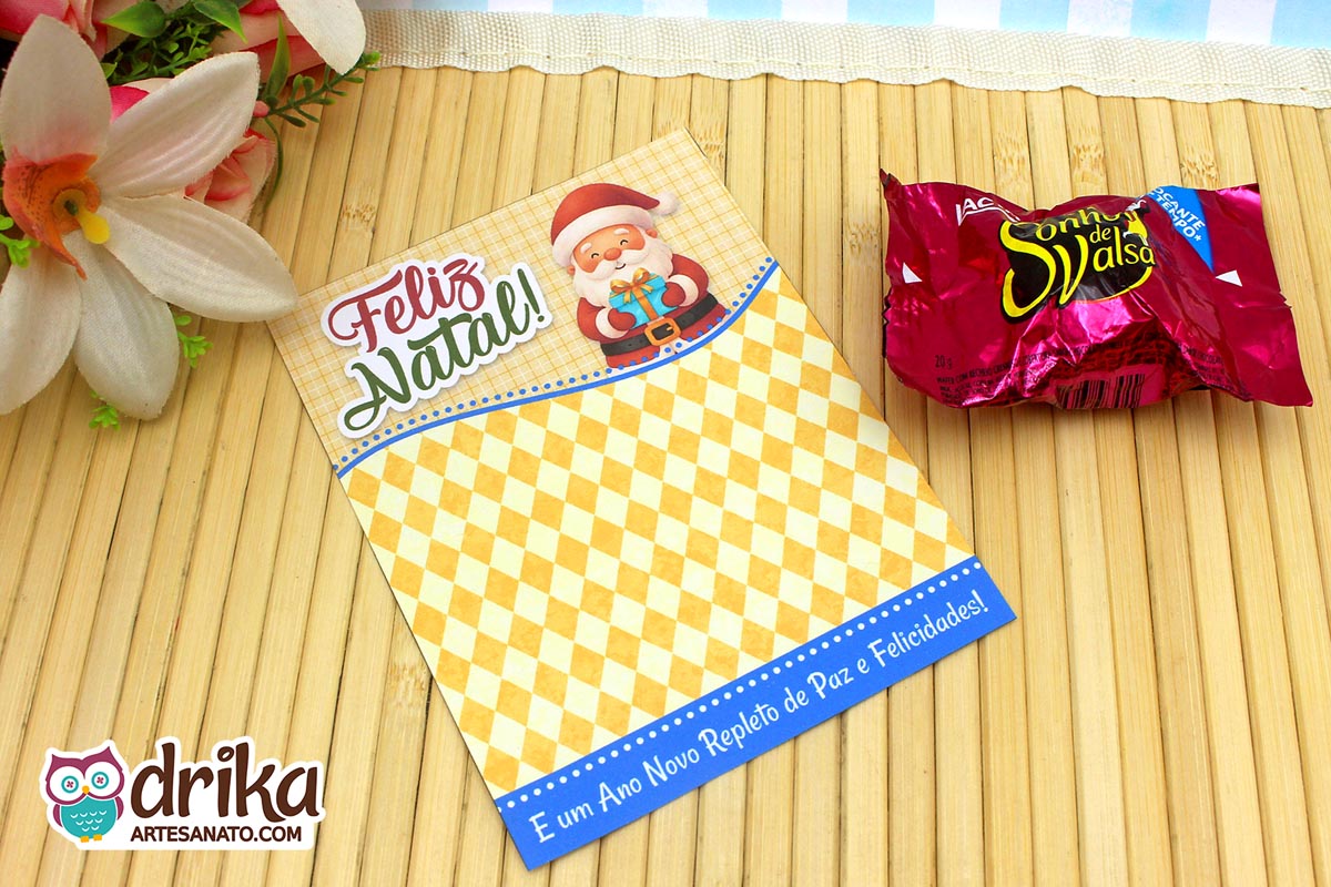 Cartão porta-bombom de Natal com Papai Noel segurando presente azul, fundo xadrez amarelo e mensagem de Boas Festas.
