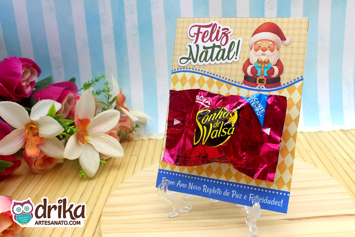 Cartão porta-bombom de Papai Noel com bombom Sonho de Valsa afixado, em frente a flores e fundo azul listrado.