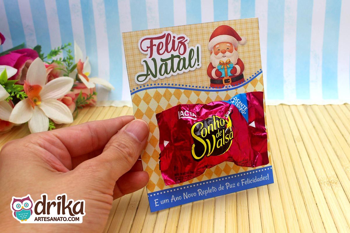 Mão segurando cartão porta-bombom de Papai Noel com bombom Sonho de Valsa, em frente a flores e fundo azul.