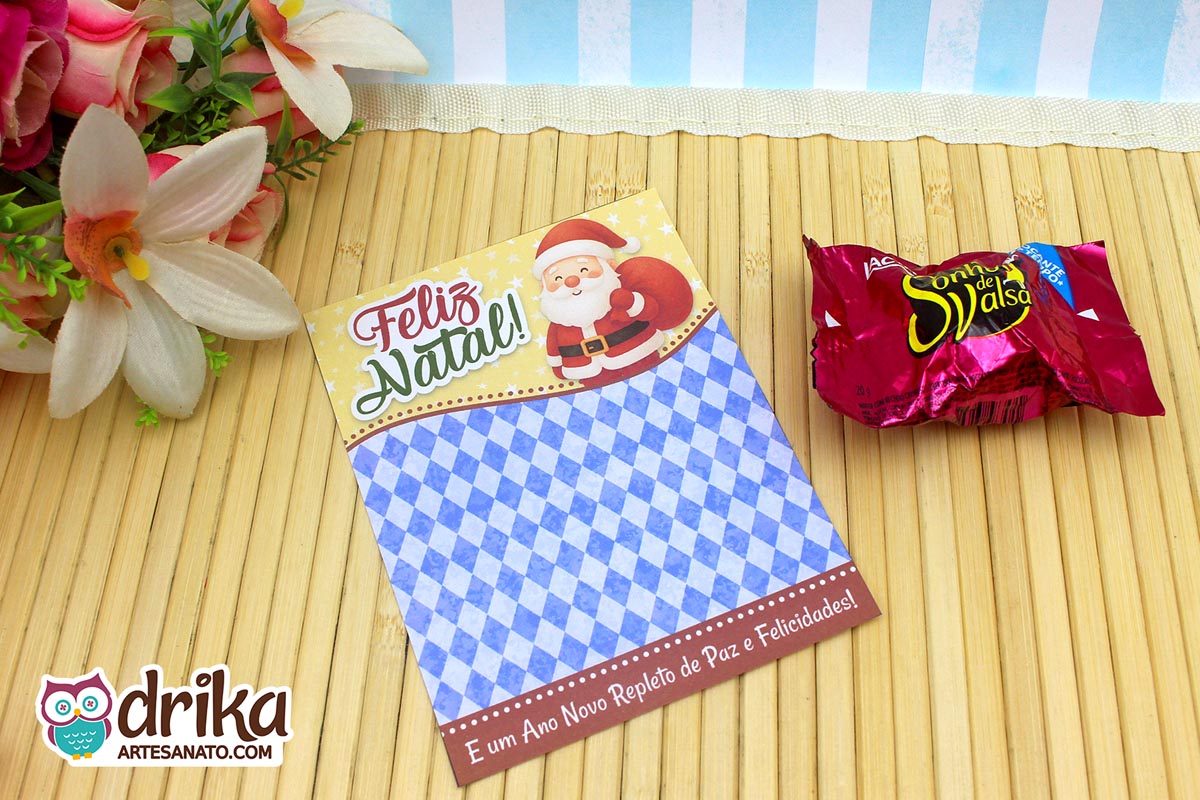 Cartão porta-bombom de Papai Noel com fundo amarelo estrelado e losangos azuis, ao lado de bombom e flores artificiais.