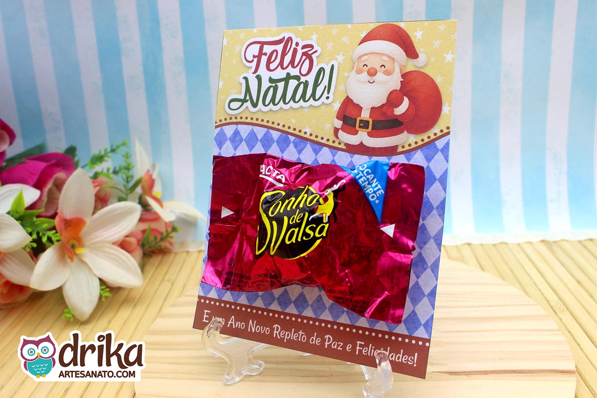Cartão porta-bombom de Papai Noel com bombom Sonho de Valsa afixado, em frente a flores artificiais.