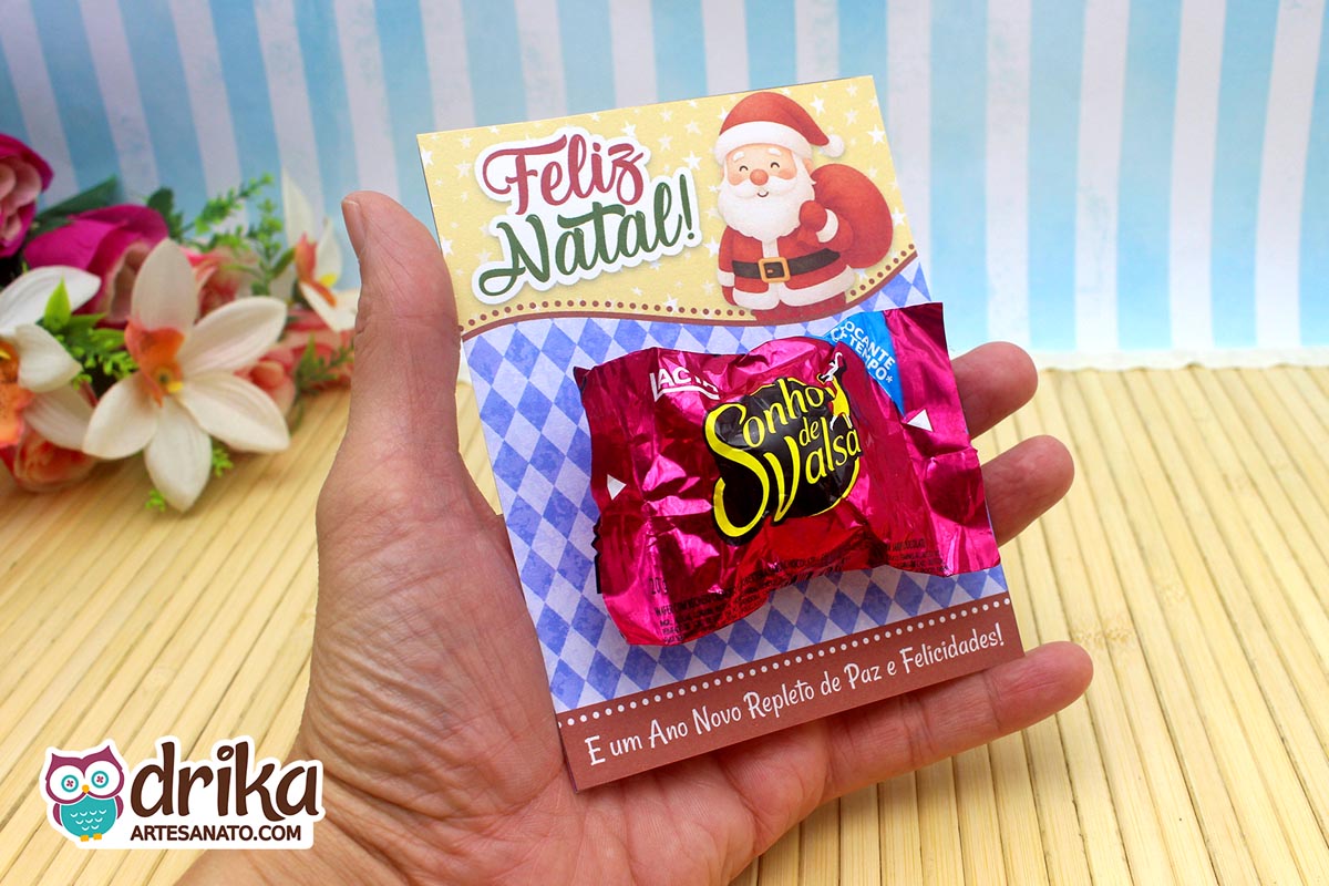 Mão segurando cartão porta-bombom de Papai Noel com bombom, com flores artificiais ao fundo.