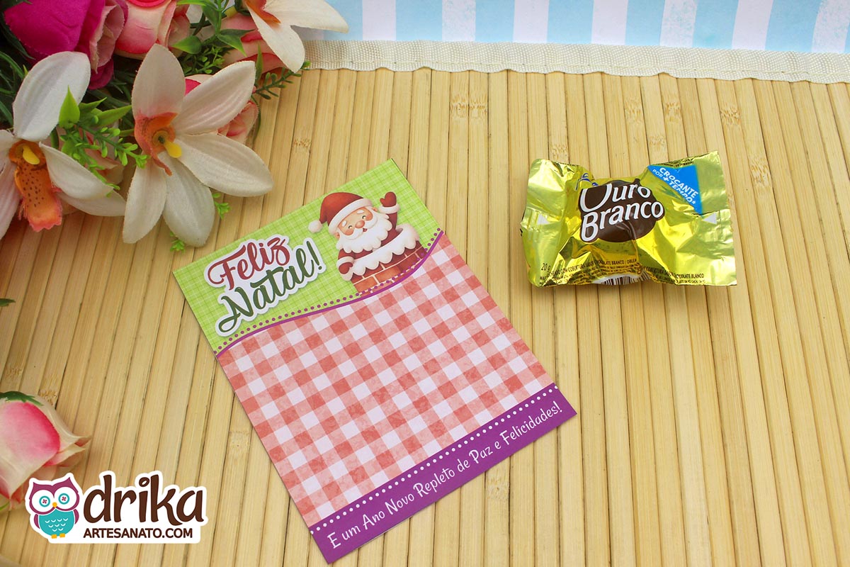 Cartão porta-bombom de Natal com Papai Noel saindo da chaminé, fundo quadriculado e bombom Ouro Branco ao lado em mesa de bambu