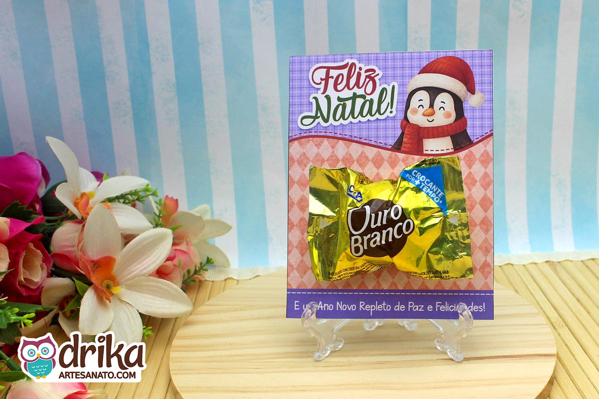 Cartão porta-bombom de pinguim com bombom Ouro Branco encaixado, em base transparente e fundo azul listrado.