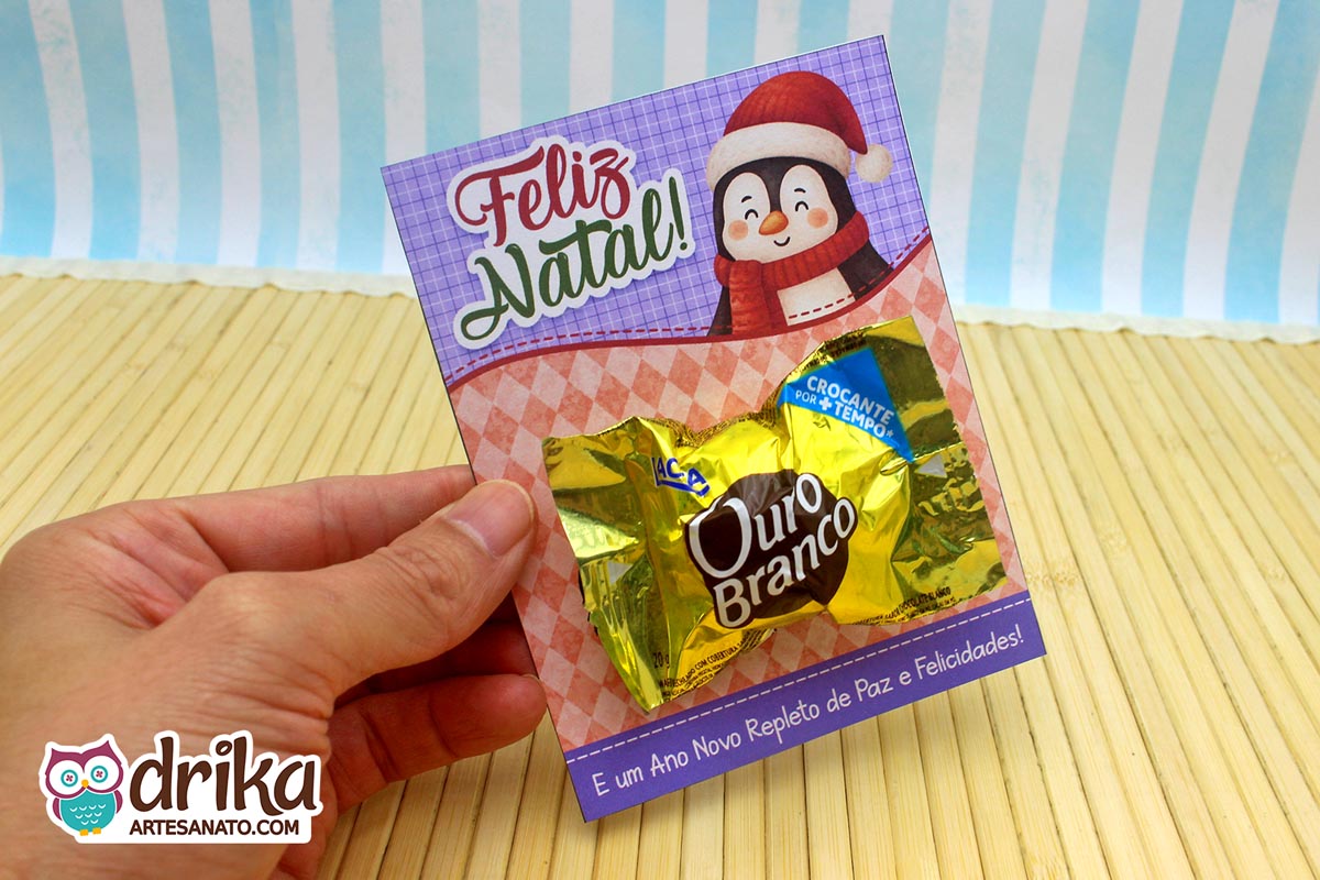 Mão segurando cartão porta-bombom de pinguim com bombom Ouro Branco em fundo de madeira e listras azuis.