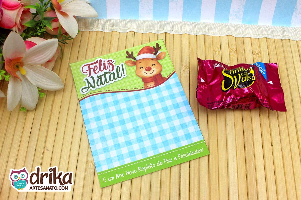 Cartão porta-bombom de Natal com rena fofa de gorro e cachecol, fundo verde xadrez azul e bombom Sonho de Valsa ao lado.