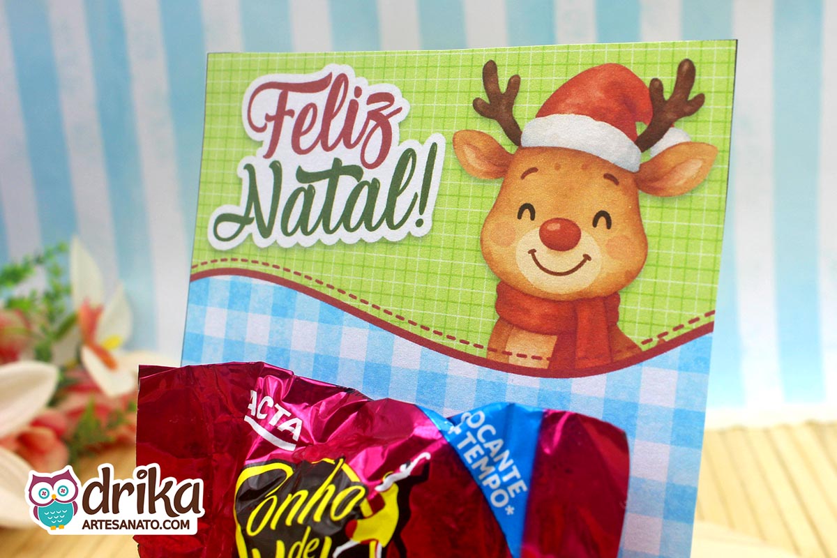 Close-up do cartão porta-bombom de rena com gorro e cachecol, com bombom Sonho de Valsa encaixado na parte inferior.