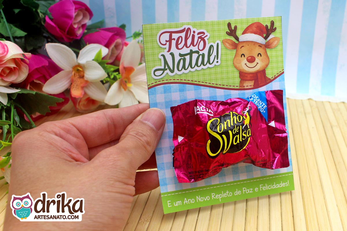 Mão segurando cartão porta-bombom de rena com bombom Sonho de Valsa, fundo verde xadrez azul e mensagem de Feliz Natal.