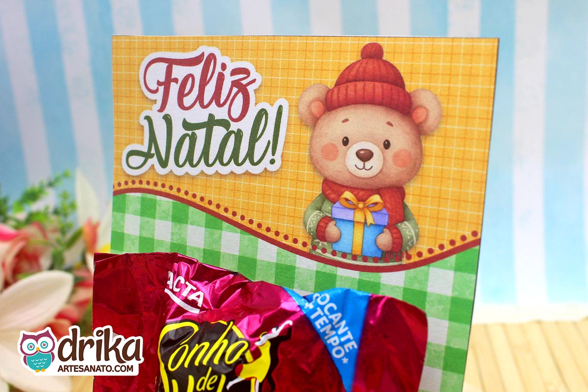 Close do cartão porta-bombom de Natal com urso de gorro e cachecol, segurando presente azul.