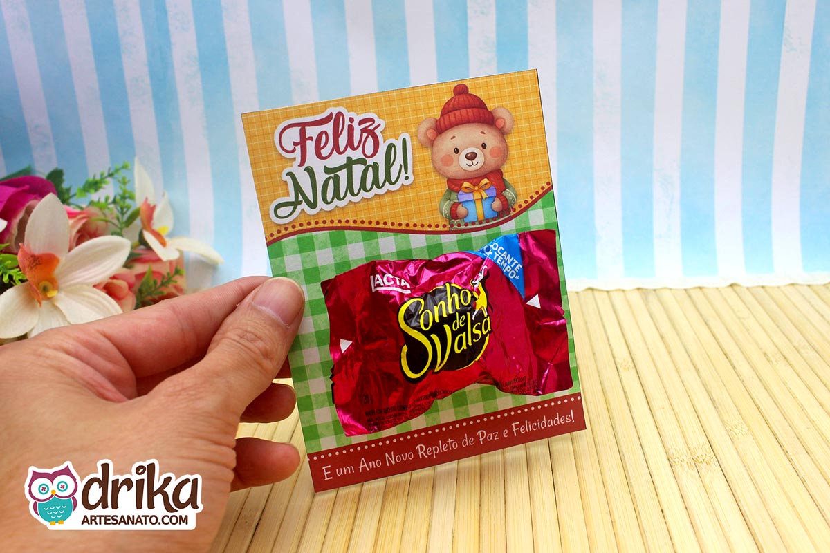 Mão segurando cartão porta-bombom de Natal com urso e bombom Sonho de Valsa fixado.