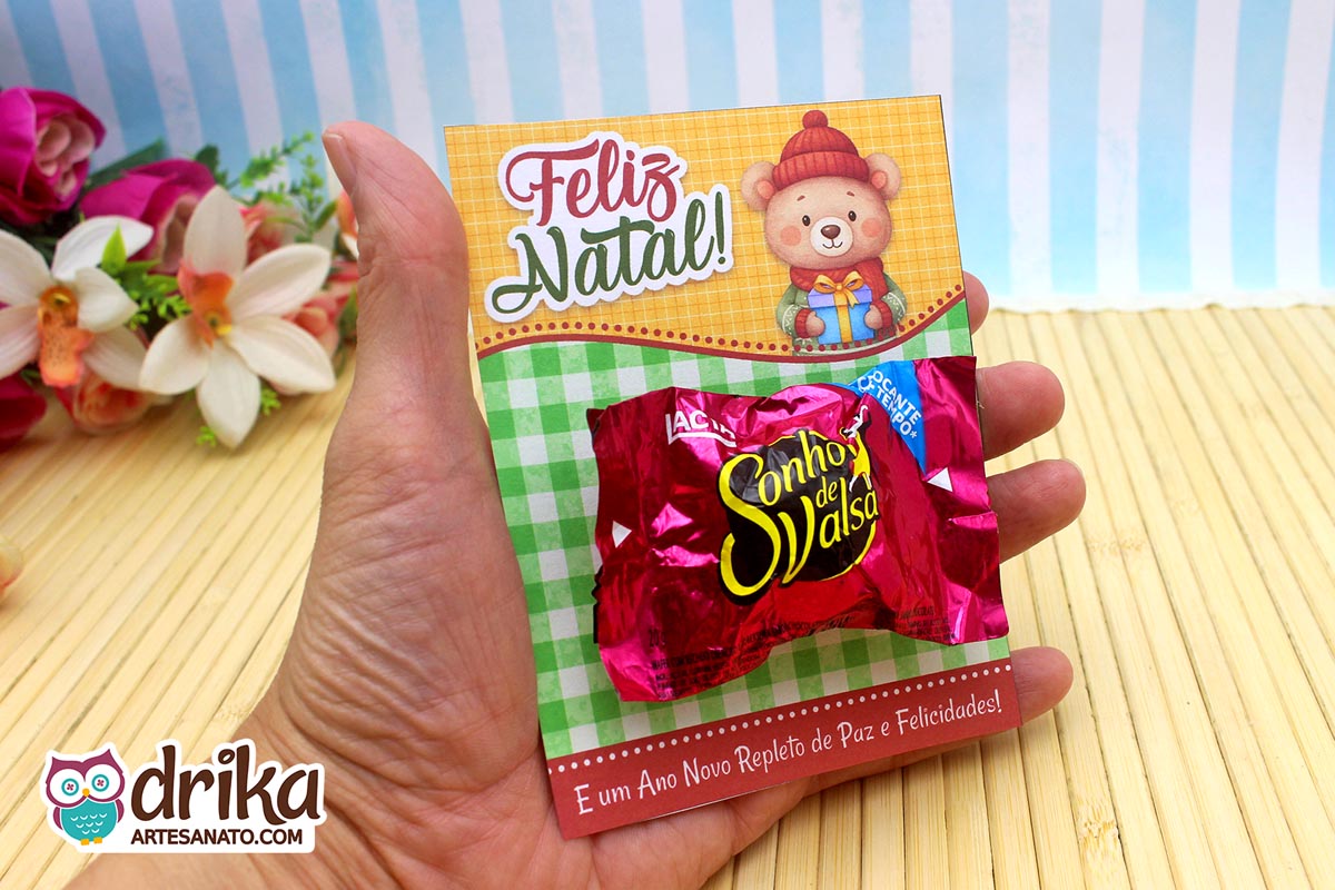 Mão segurando cartão porta-bombom de Natal com urso e bombom Sonho de Valsa fixado.