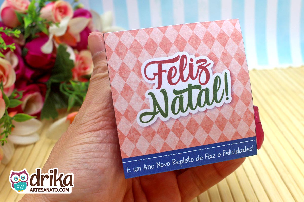 Mão segurando porta-bombom de Natal com estampa geométrica rosa e mensagem de Feliz Natal.