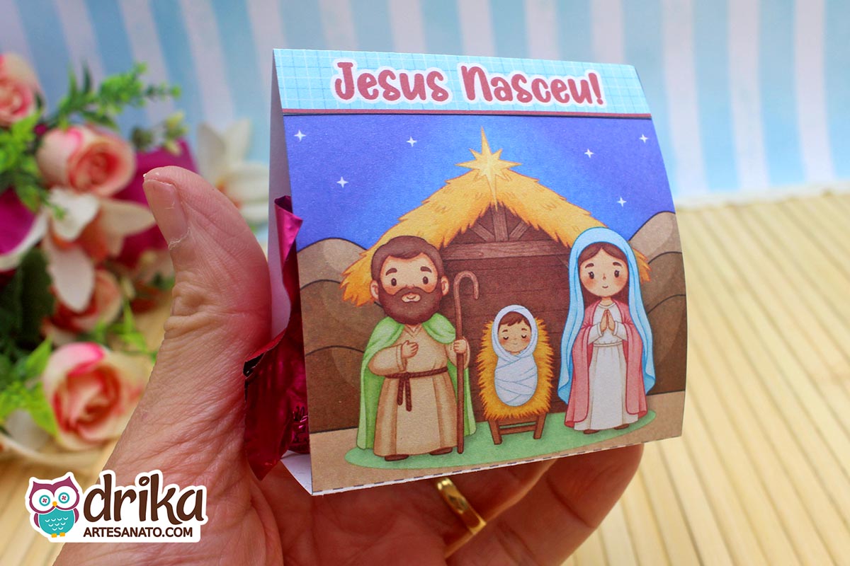 Mão segurando porta-bombom de Natal com ilustração do presépio e a frase "Jesus Nasceu!" em fundo azul.