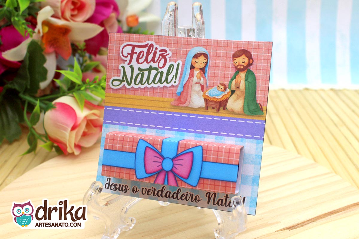 Cartão porta-bis de Natal com ilustração da Sagrada Família e chocolate Bis embalado, em suporte transparente sobre mesa de madeira.