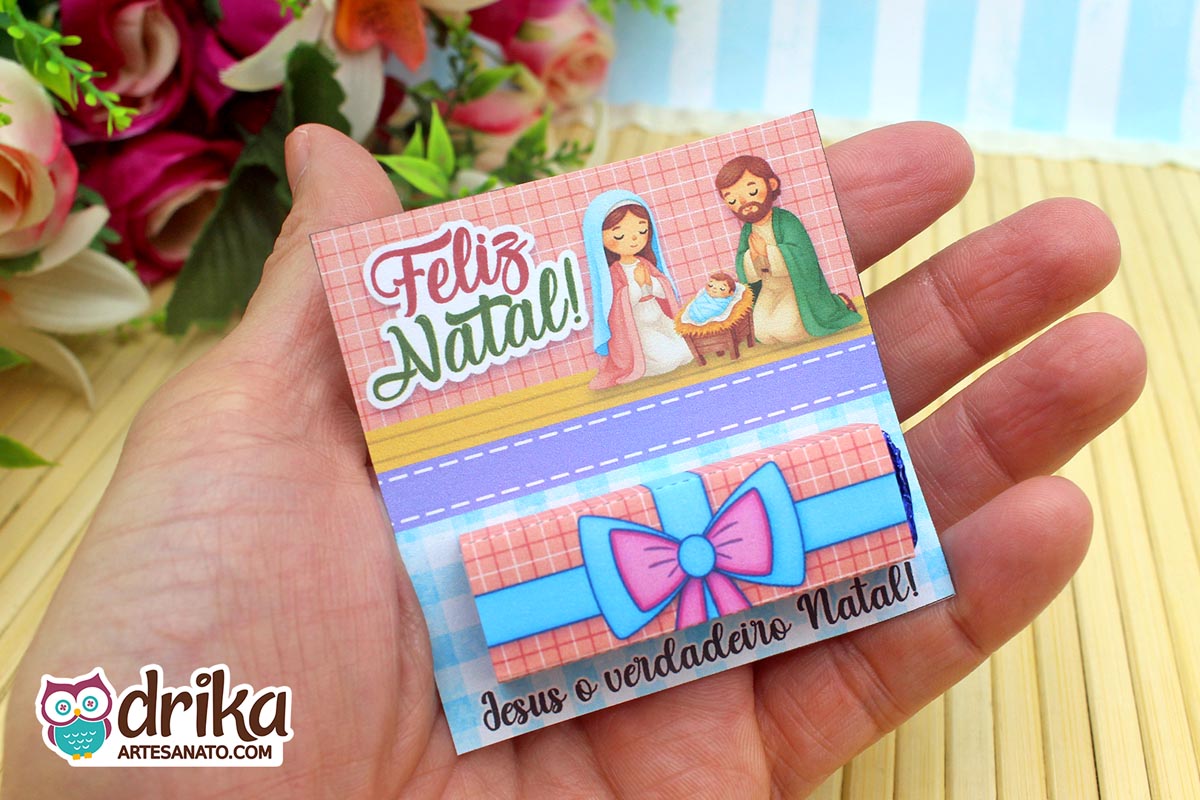 Mão segurando cartão porta-bis de Natal com ilustração da Sagrada Família e chocolate Bis embalado, com fundo de flores e bambu.