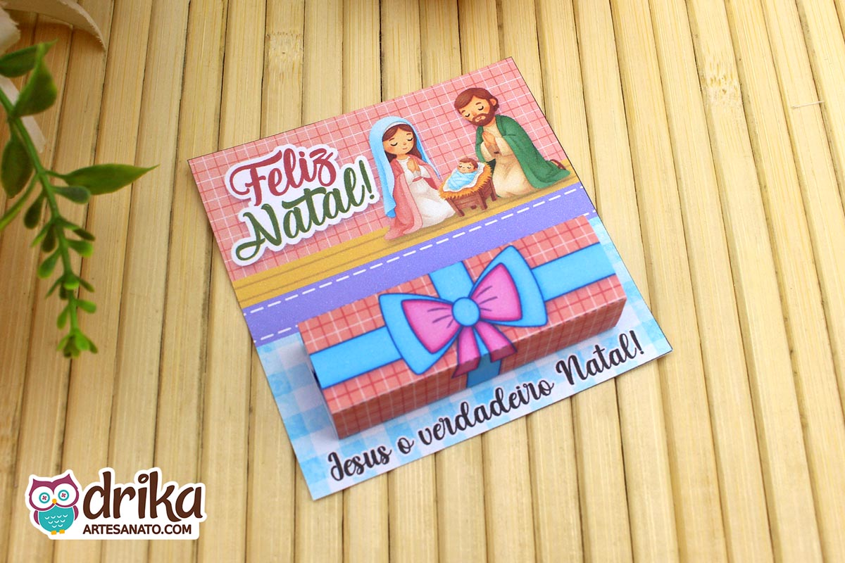 Cartão porta-bis de Natal com ilustração da Sagrada Família e chocolate Bis embalado, sobre fundo de bambu e folhagem verde.