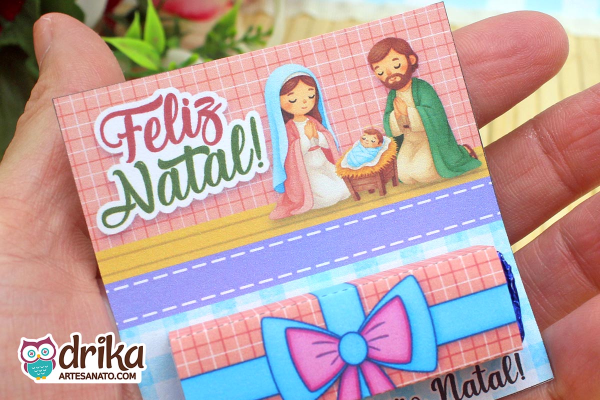 Close-up de mão segurando cartão porta-bis de Natal com ilustração da Sagrada Família e chocolate Bis embalado, em fundo desfocado.