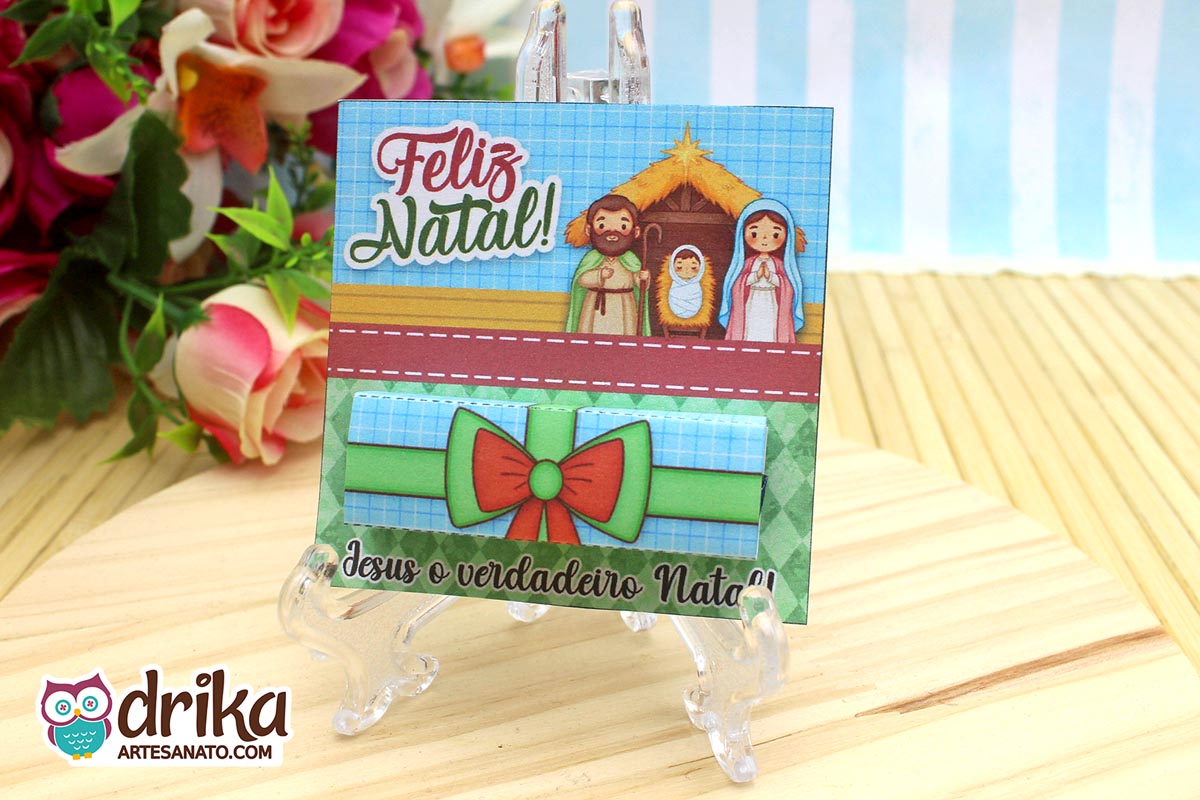 Cartão porta-bis de Natal com ilustração da Sagrada Família em suporte transparente sobre mesa de madeira.