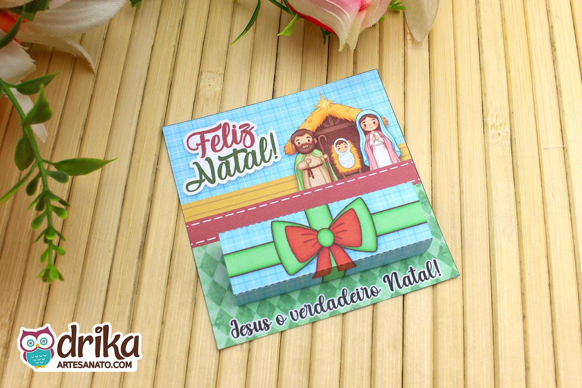 Cartão porta-bis de Natal com ilustração da Sagrada Família e laço verde e vermelho sobre mesa de bambu.