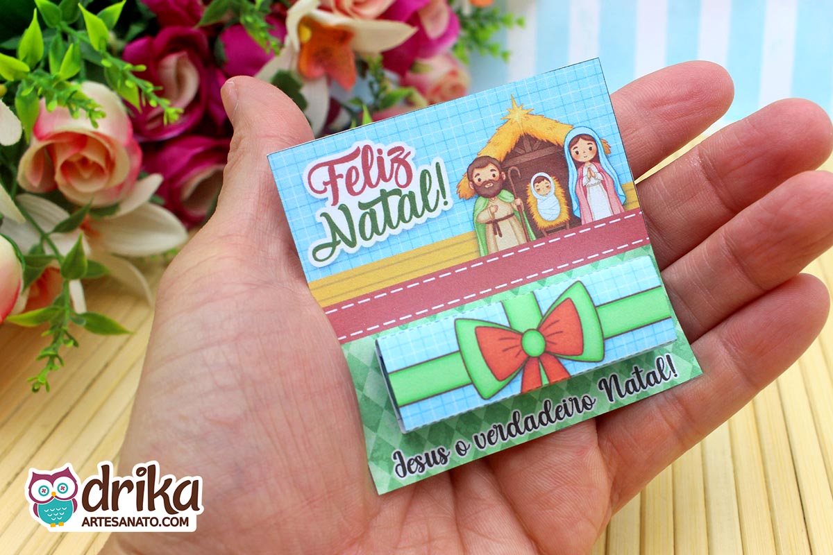 Mão segurando cartão porta-bis de Natal com ilustração da Sagrada Família e laço verde e vermelho.