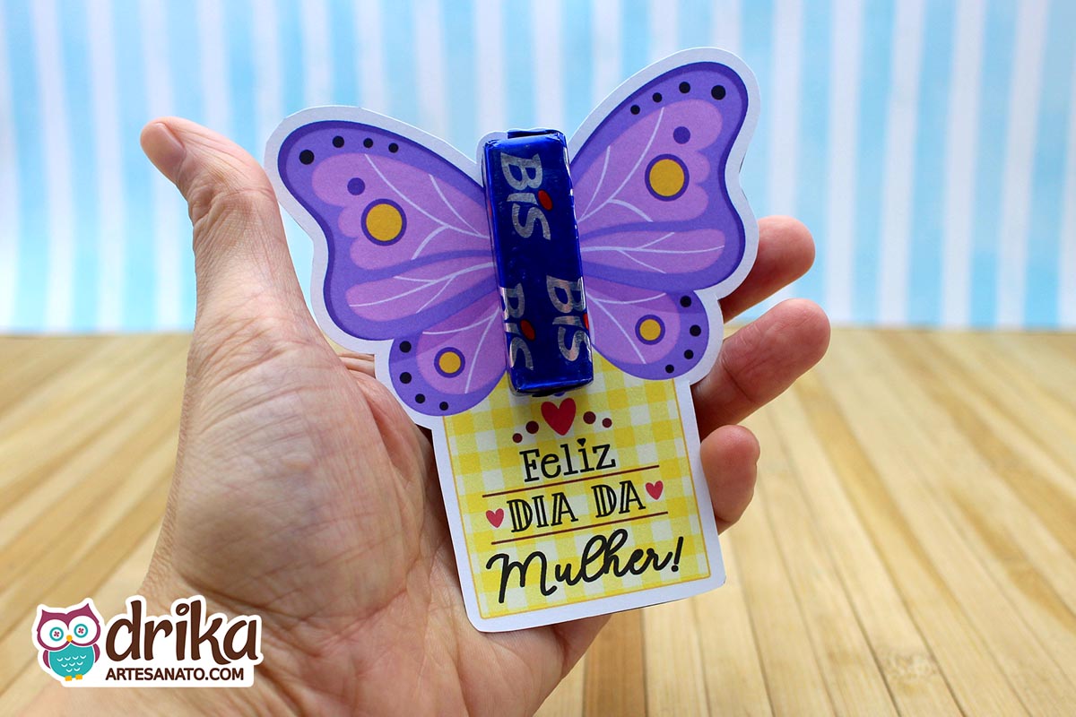 Mão segurando cartão porta-bis de borboleta roxa com detalhes amarelos e brancos, e mensagem "Feliz Dia da Mulher"