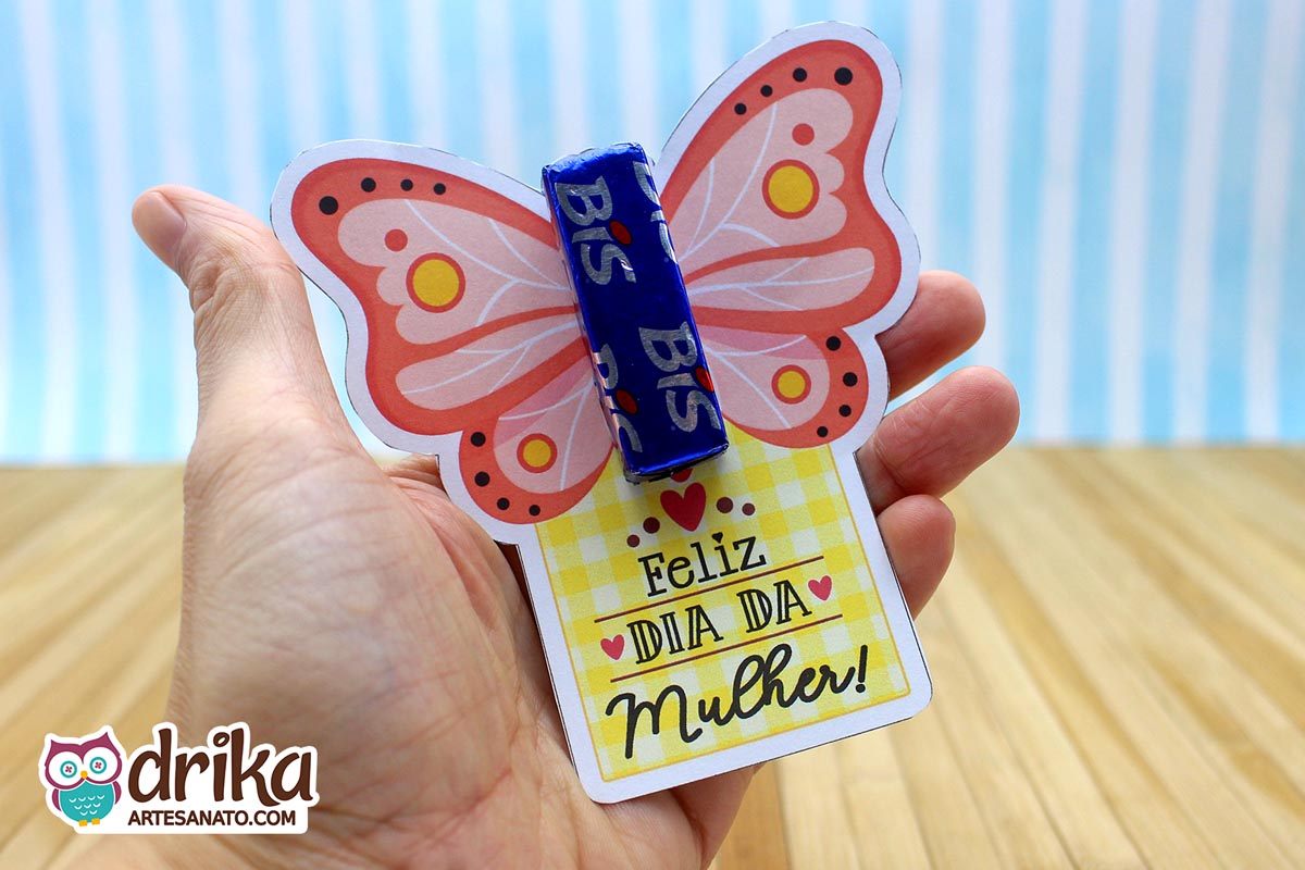 Mão segurando cartão porta-bis de borboleta coral com detalhes amarelos e brancos, e mensagem "Feliz Dia da Mulher"