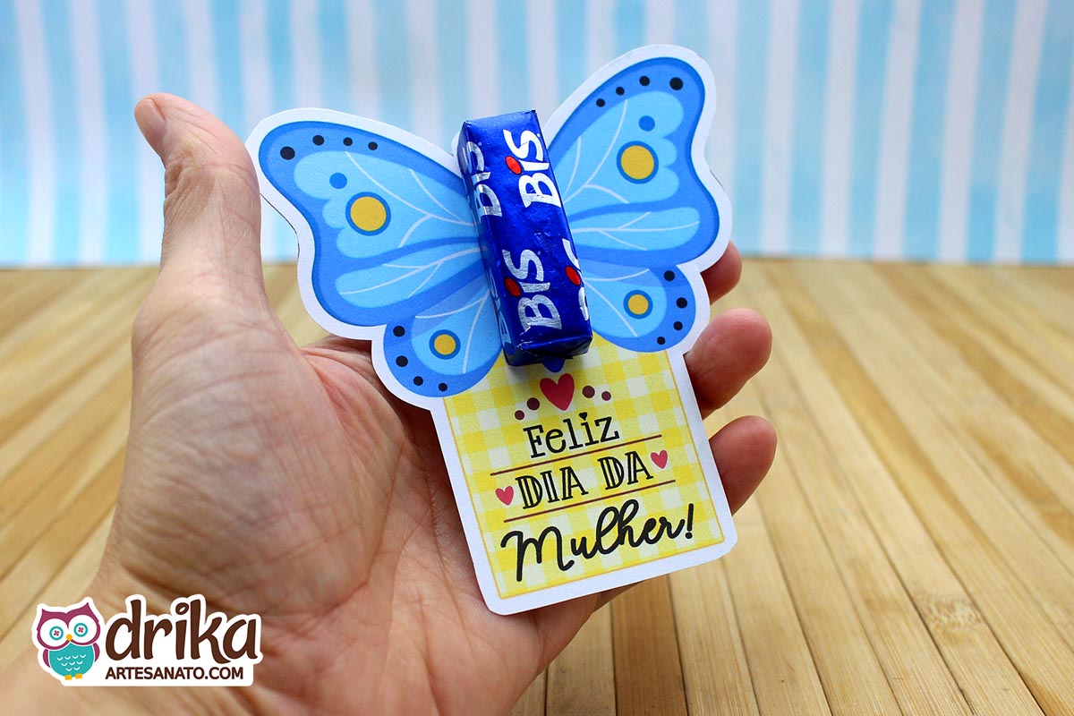 Mão segurando cartão porta-bis de borboleta azul com detalhes amarelos e brancos, e mensagem "Feliz Dia da Mulher"