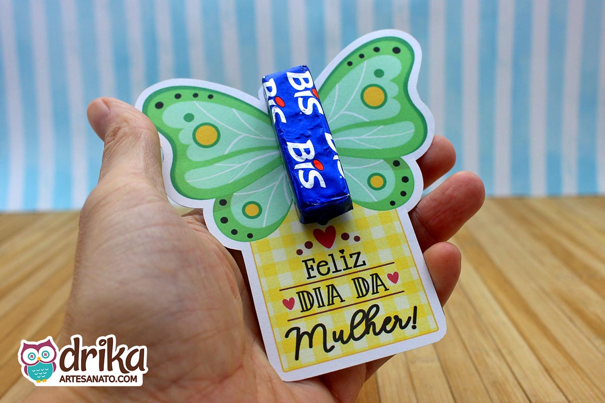 Mão segurando cartão porta-bis de borboleta verde com detalhes amarelos e brancos, e mensagem "Feliz Dia da Mulher"