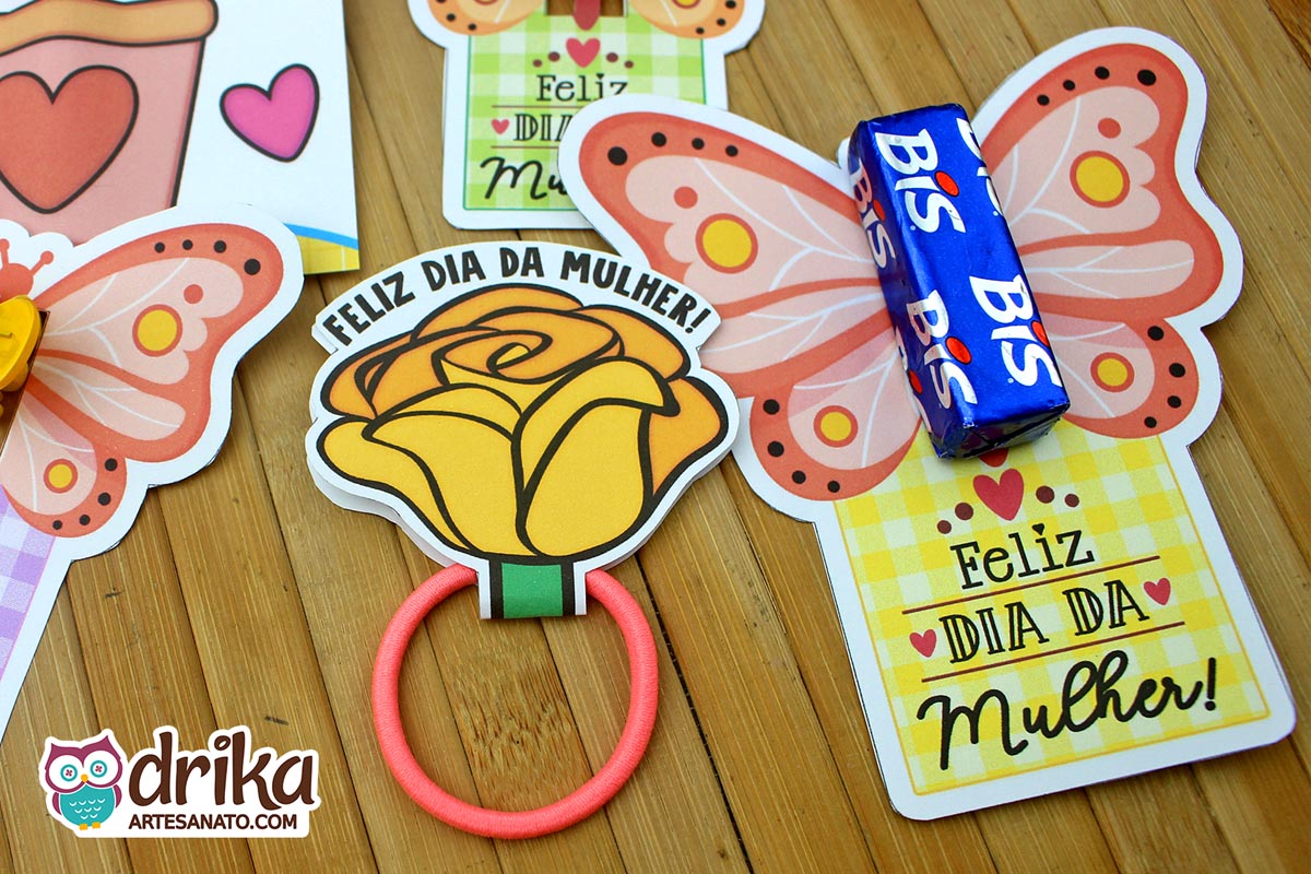 Três lembrancinhas de Dia da Mulher sobre mesa de madeira: borboleta coral com Bis, rosa amarela com elástico de cabelo rosa