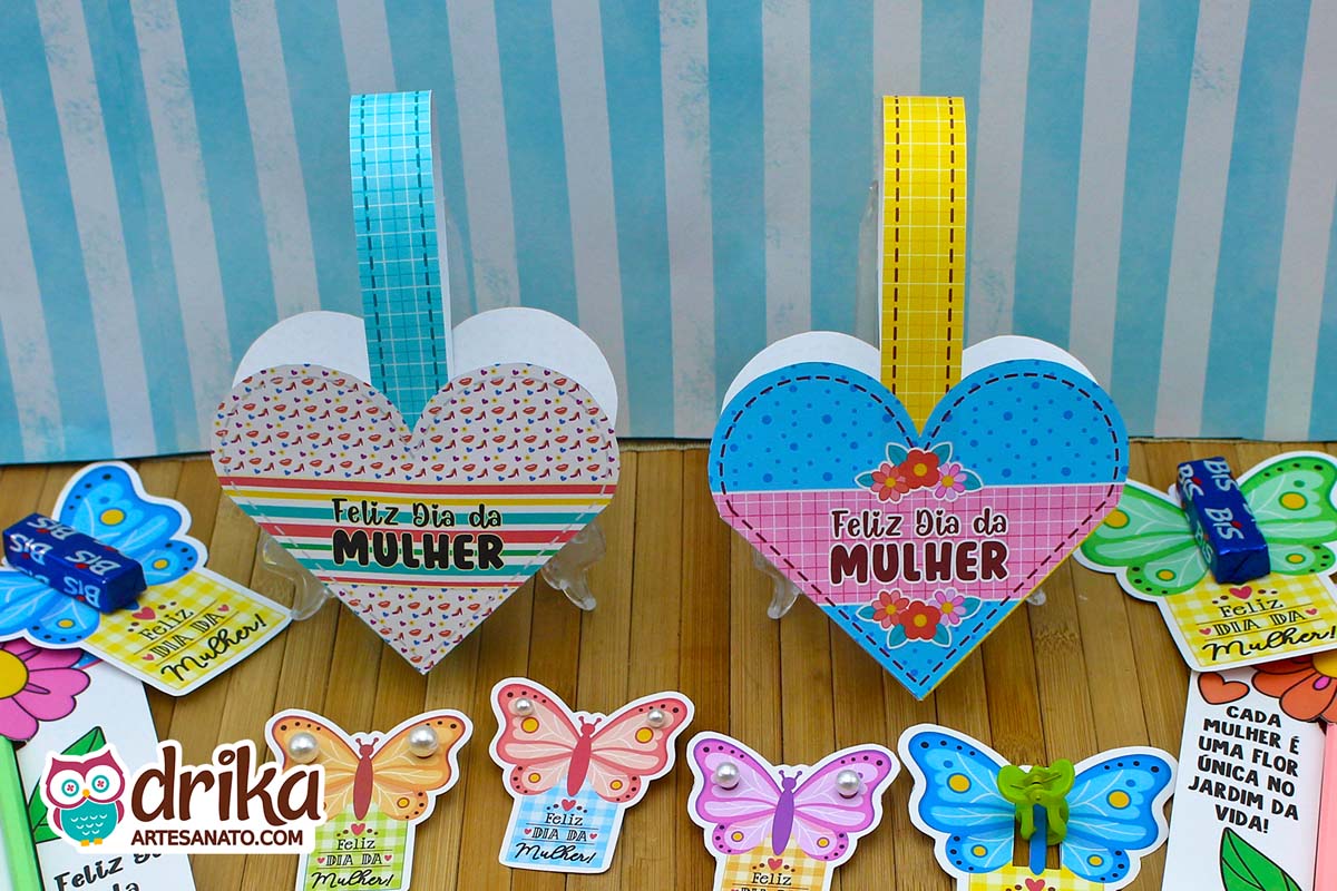 Duas cestinhas de coração de papel, uma com estampa de corações e outra azul de bolinhas e flores, com alças e a frase "Feliz Dia da Mulher"