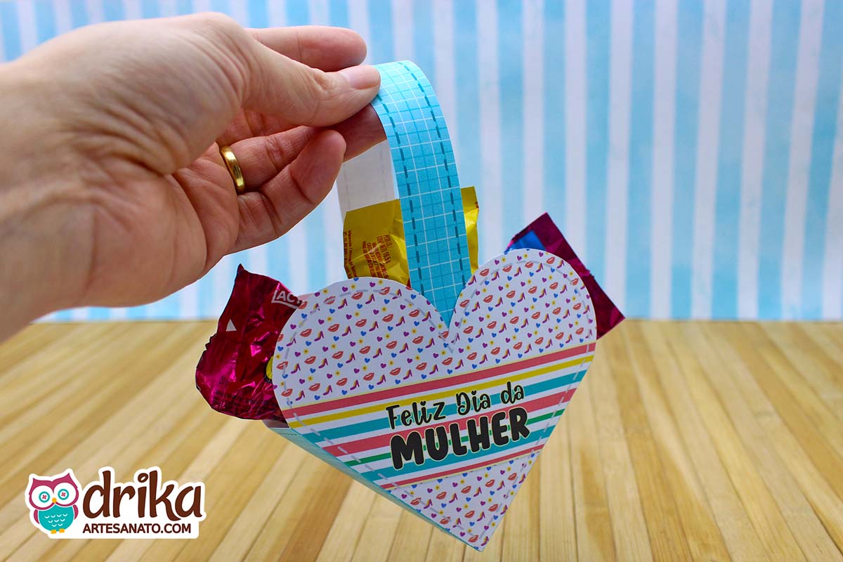 Mão segurando pela alça uma cestinha de coração de papel com estampa de sapatinhos e listras, recheada com bombons