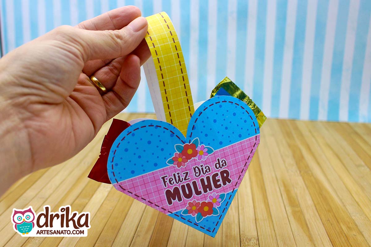 Mão segurando pela alça uma cestinha de coração de papel azul com bolinhas e flores, recheada com bombons