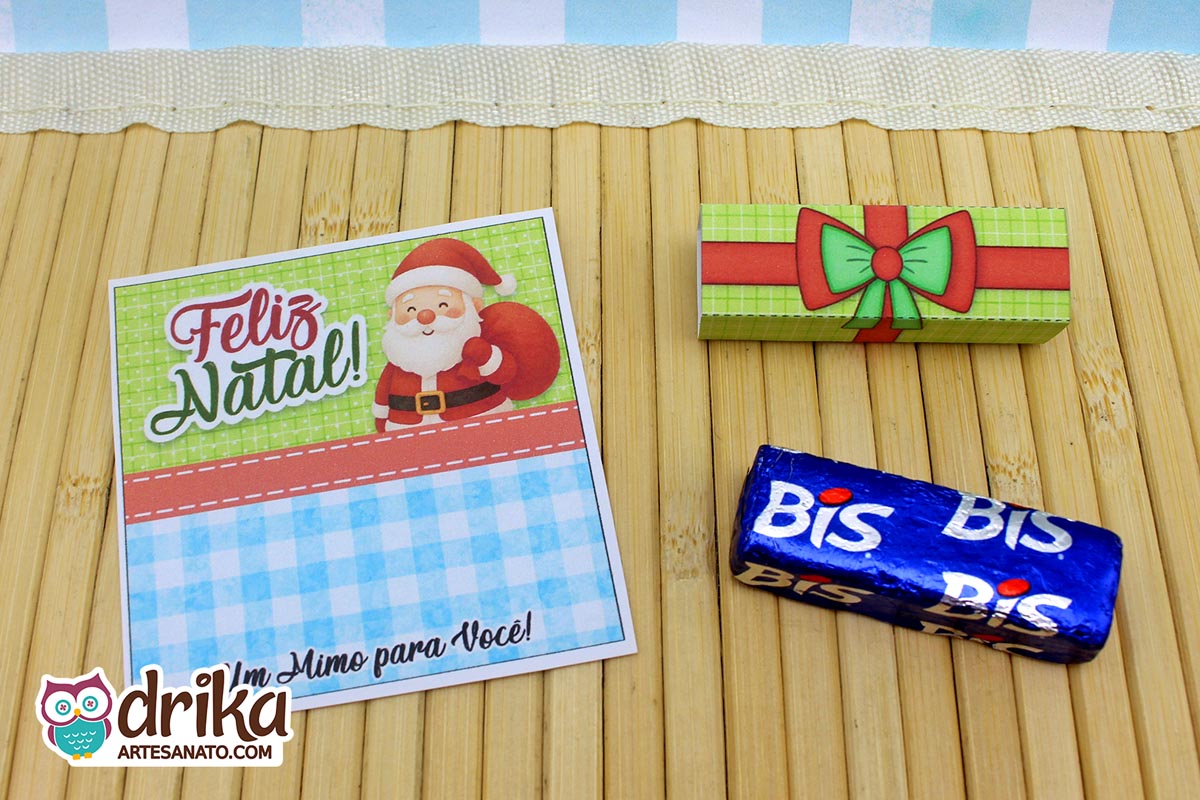 Cartão de Natal com Papai Noel e "Feliz Natal", cinta decorada de presente e chocolate Bis separados em mesa de bambu