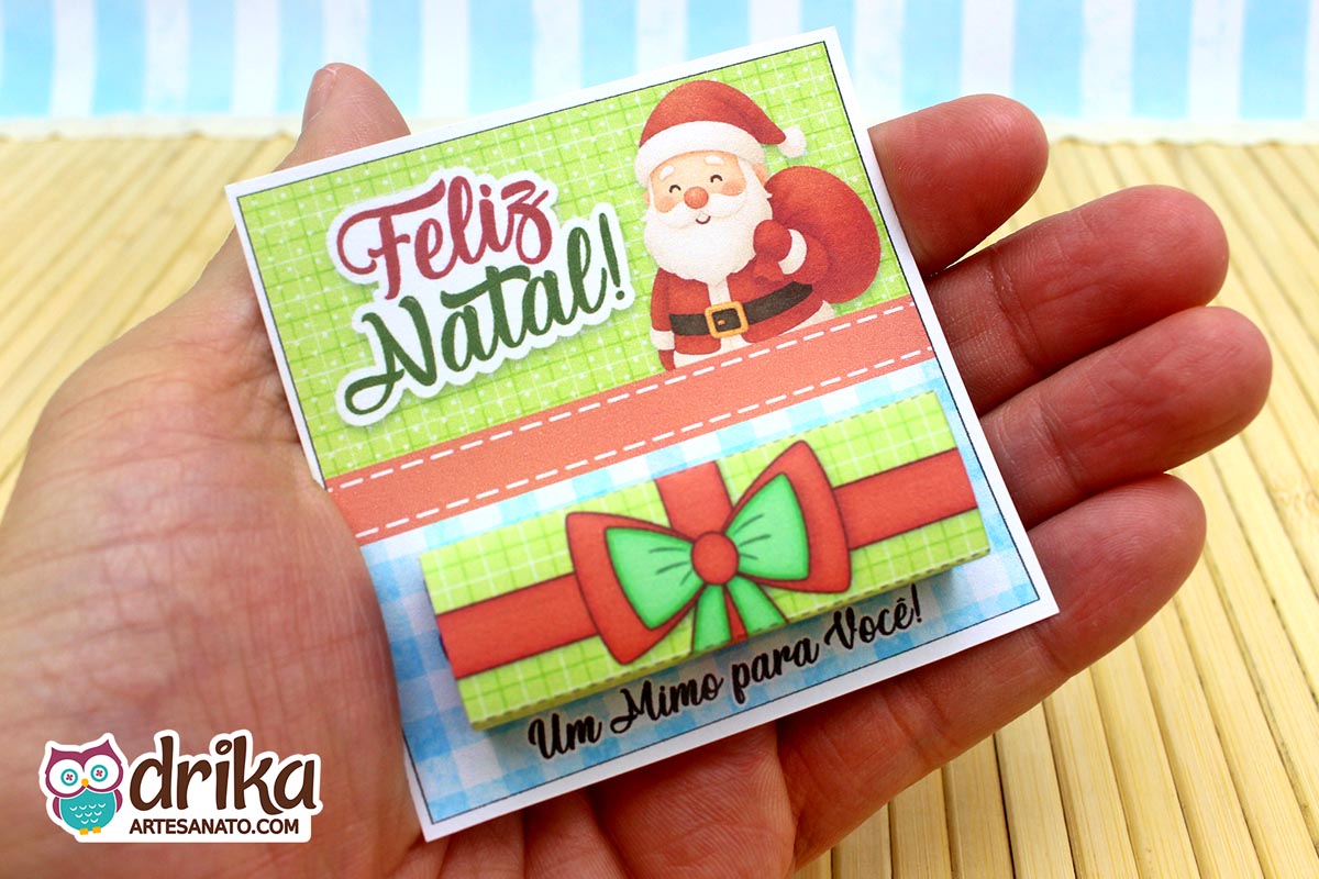 Mão segurando cartão porta-bis de Papai Noel montado, com chocolate Bis encaixado na cinta de presente e "Feliz Natal"