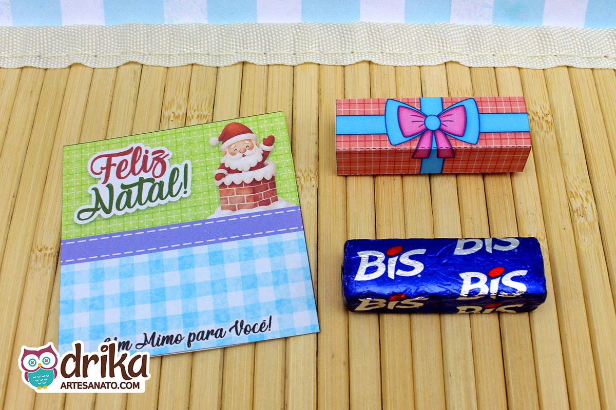 Cartão porta-bis de Papai Noel na chaminé, cinta de presente com laço pink e azul, e um chocolate Bis azul, sobre bambu