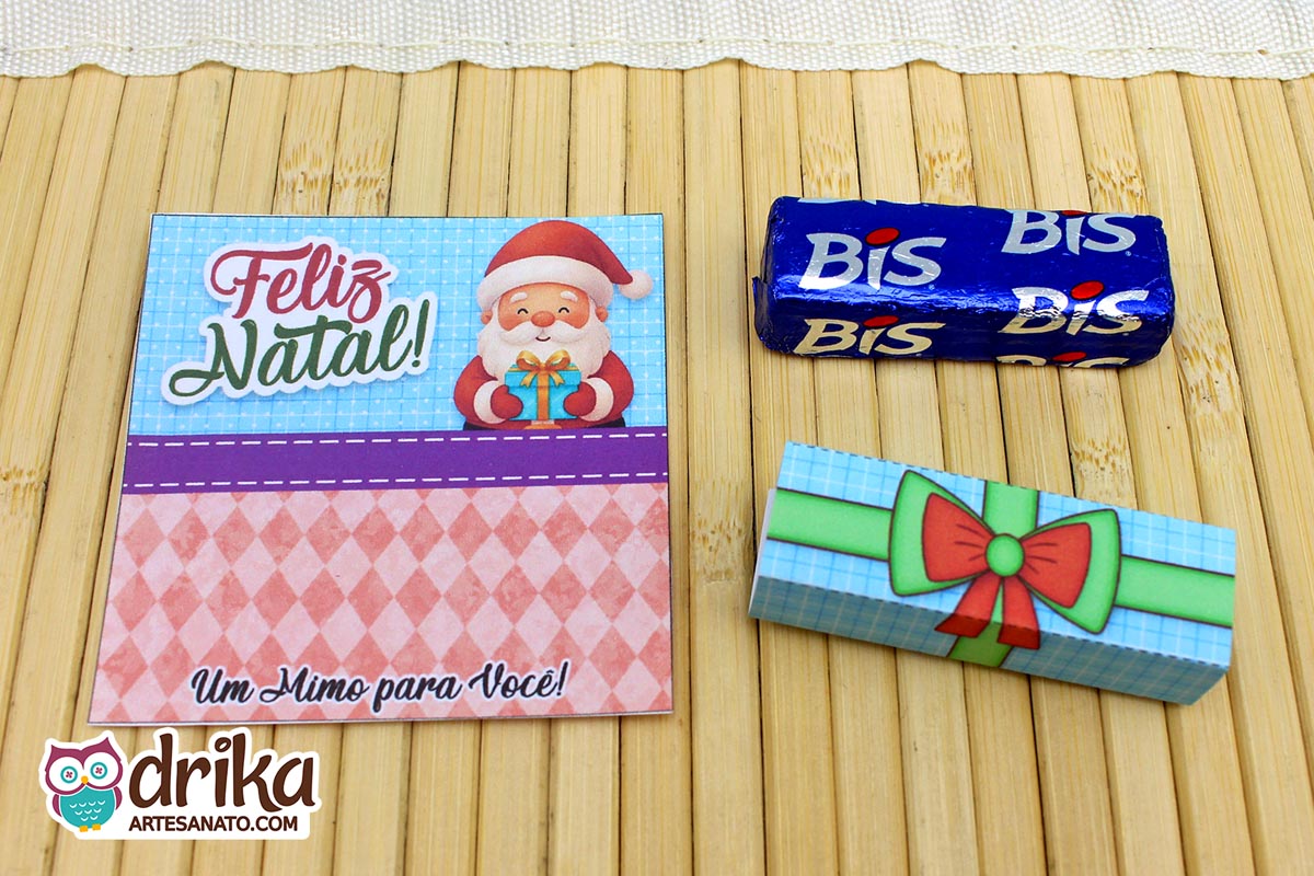 Cartão porta-bis de Papai Noel com "Feliz Natal", cinta verde de presente e chocolate Bis azul separados em esteira de bambu