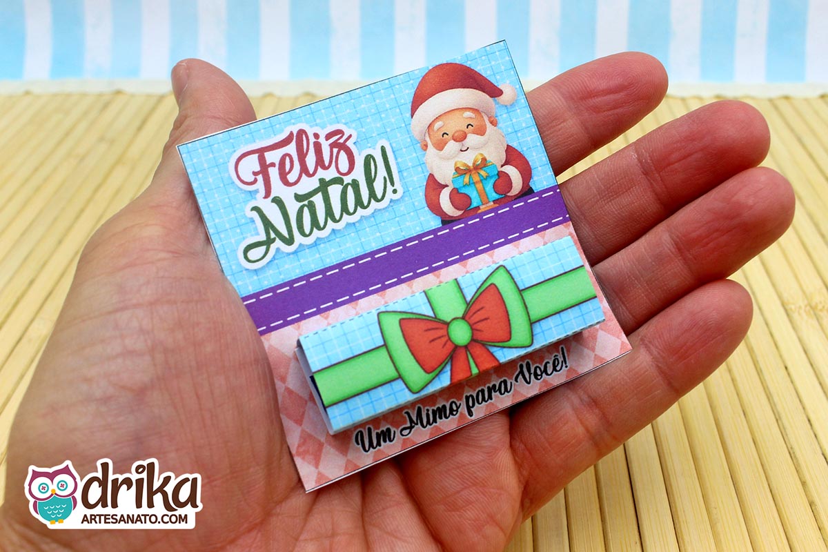 Mão segurando um cartão porta-bis de Papai Noel montado, com chocolate Bis embrulhado em cinta de presente verde