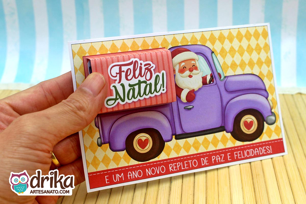 Mão segurando cartão porta-bis de Papai Noel em caminhonete lilás, com cinta "Feliz Natal" já encaixada na caçamba