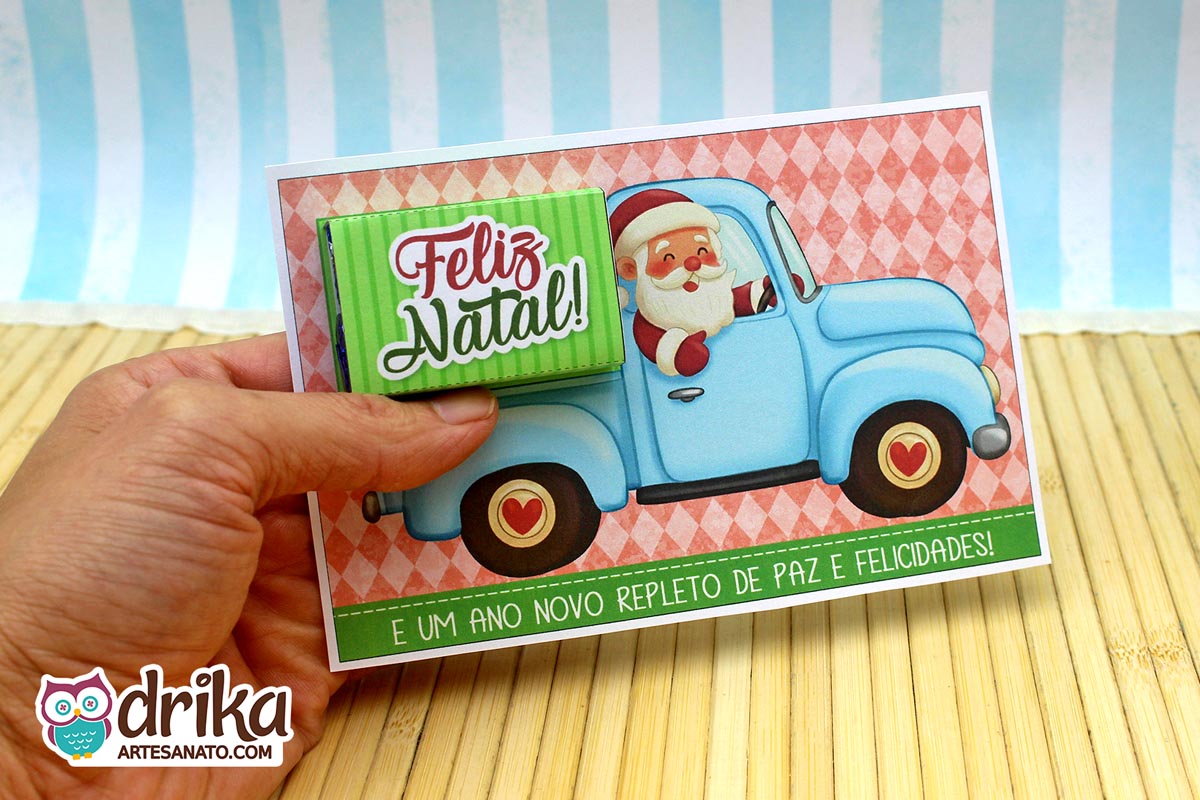 Mão segurando cartão porta-bis de Papai Noel em caminhonete azul clara, com cinta "Feliz Natal" já encaixada