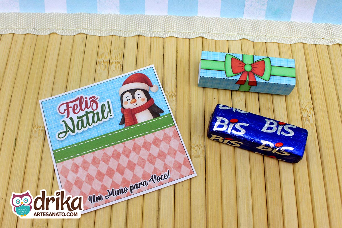 Cartão de Natal com pinguim de gorro e cachecol, cinta de presente azul com laço vermelho e chocolate Bis azul em mesa de bambu