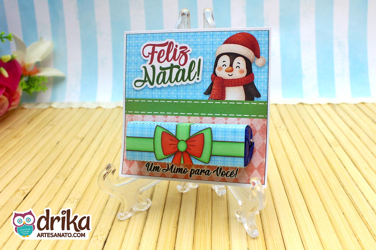 Cartão porta-bis de pinguim com gorro e cachecol, chocolate Bis na cinta de presente azul com laço vermelho, em suporte