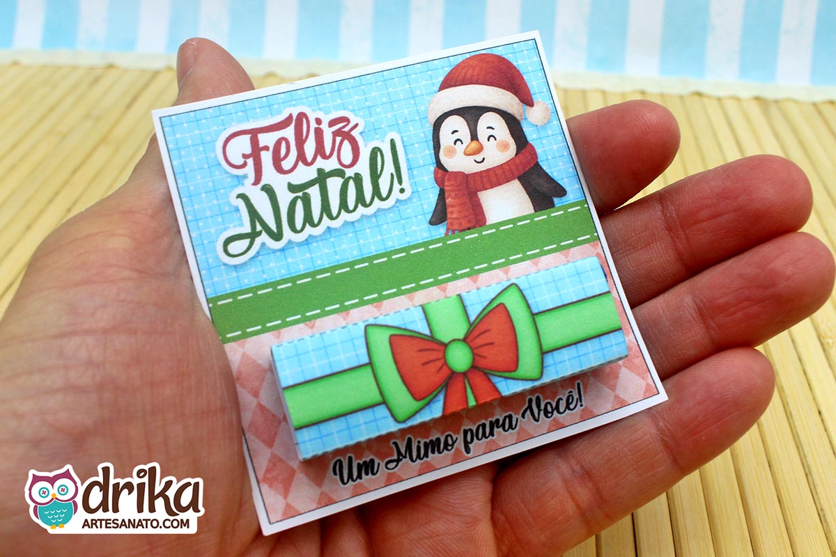 Mão segurando cartão porta-bis de pinguim com gorro e cachecol, chocolate Bis na cinta de presente azul com laço vermelho