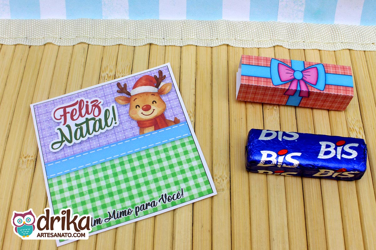 Cartão de Natal com rena fofa de gorro e cachecol, "Feliz Natal!", ao lado de um chocolate Bis e sua cinta de presente