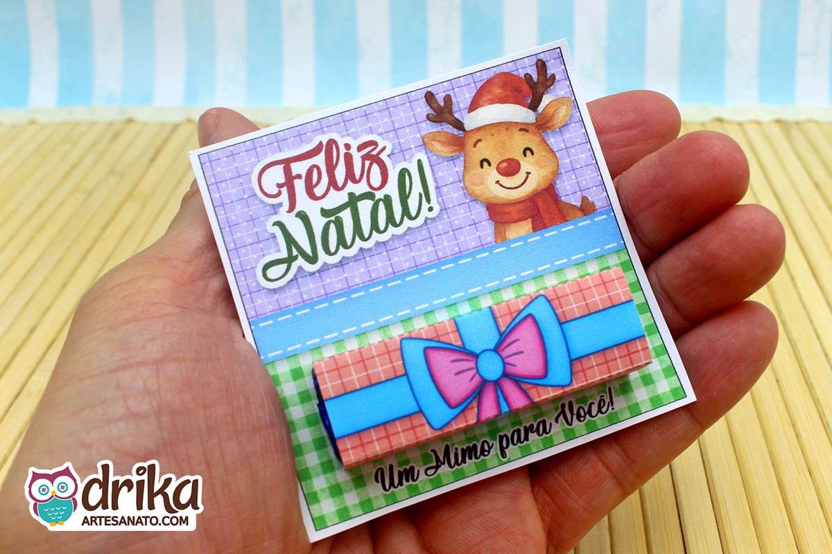Mão segurando o cartão porta-bis de Natal com rena fofa, "Feliz Natal!", e o chocolate Bis já encaixado na cinta de presente