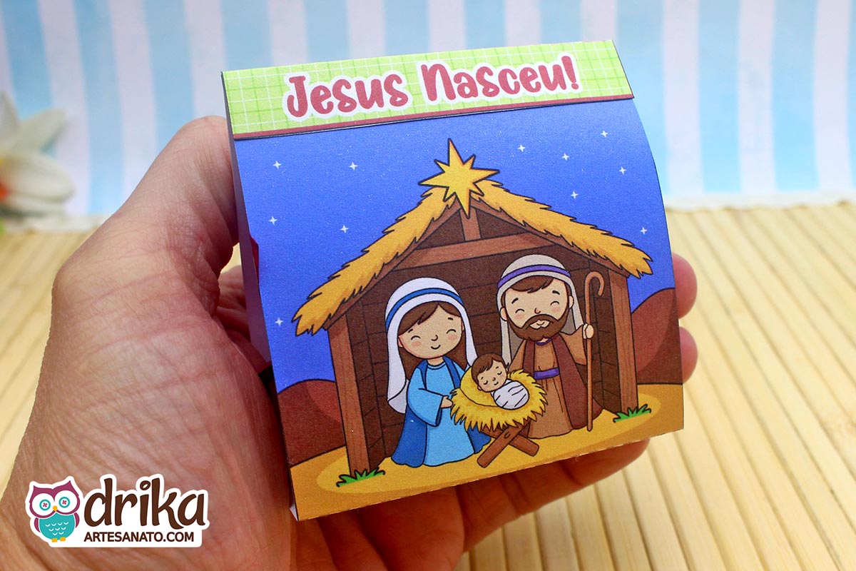 Mão segurando porta-bombom de Natal com ilustração da Sagrada Família e frase "Jesus Nasceu!" em fundo azul estrelado.