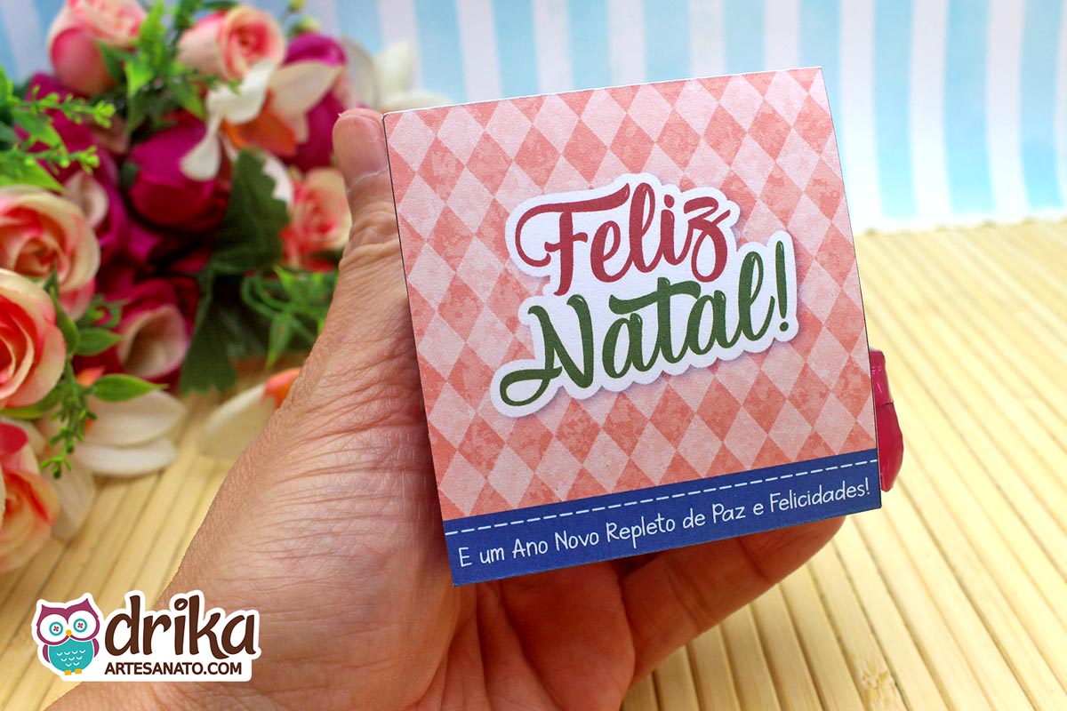 Mão segurando porta-bombom de Natal com mensagem "Feliz Natal! E um Ano Novo Repleto de Paz e Felicidades!" em fundo coral.